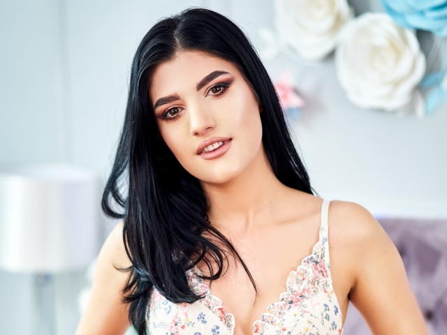 AngelaFay with long black hair, brown eyes, and floral lace bra, showing her tits, has a slight smile, and looks directly at the camera in a bright, soft-lit photo.