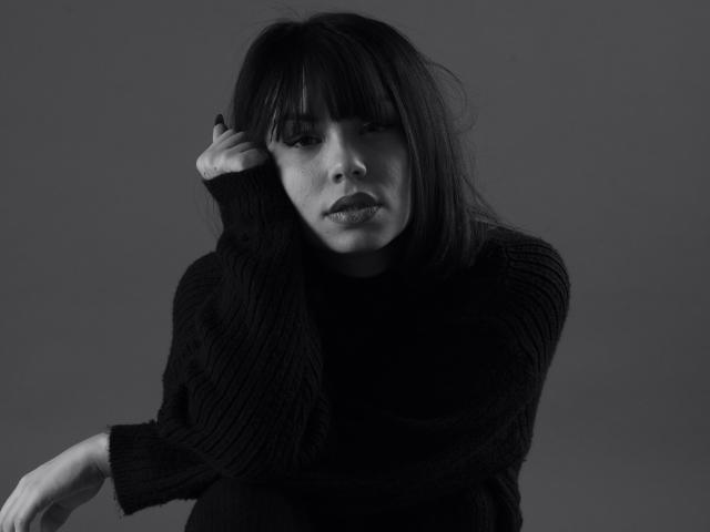 KassiKassou, con cabello oscuro y liso con flequillo, vestida con un suéter negro texturizado, mirando ligeramente hacia abajo, con la mano tocándose la cabeza, en tonos grises, con un ambiente melancólico, en blanco y negro, seria, atractiva, linda..