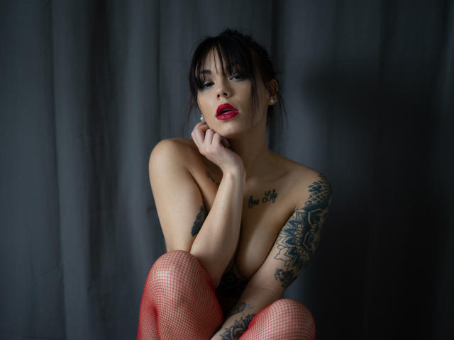 KassiKassou, com meias de rede vermelhas, tatuagens e batom vermelho, sentada sem blusa, com os braços cruzados e as mãos no queixo, olhando para a câmera. Seios visíveis. Fundo escuro. O rosto de KassiKassou está sério..