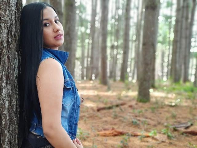 SharolXD with long black hair, wearing a blue denim sleeveless vest, leans against a tree in a forest, looking over her shoulder with a subtle smile.