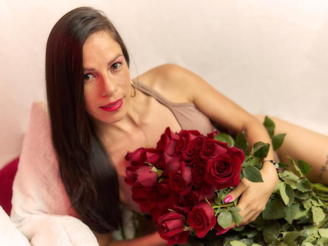 MiaKambell, con cabello castaño largo, lápiz labial rojo y un vestido color piel, sosteniendo un ramo de rosas rojas, acostada sobre un cojín rosa, sonriendo a la cámara..