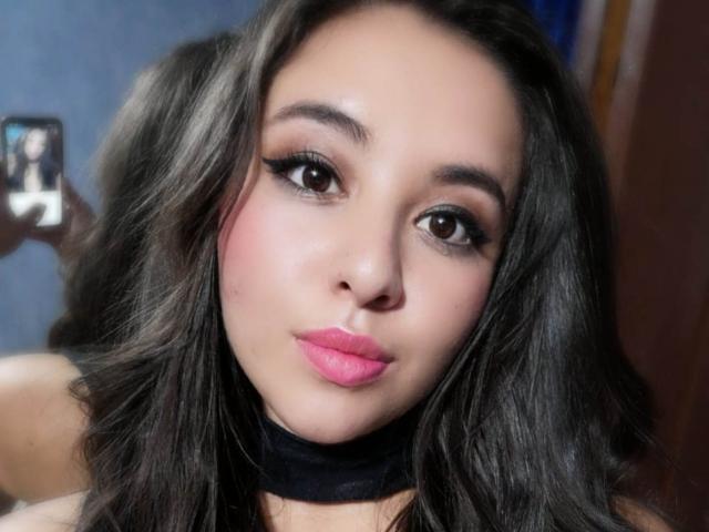 SantaRodrigues a les cheveux bruns foncés ondulés, du rouge à lèvres rose, du crayon noir pour les yeux et un collier noir. Elle tient un téléphone à sa gauche et prend un selfie..