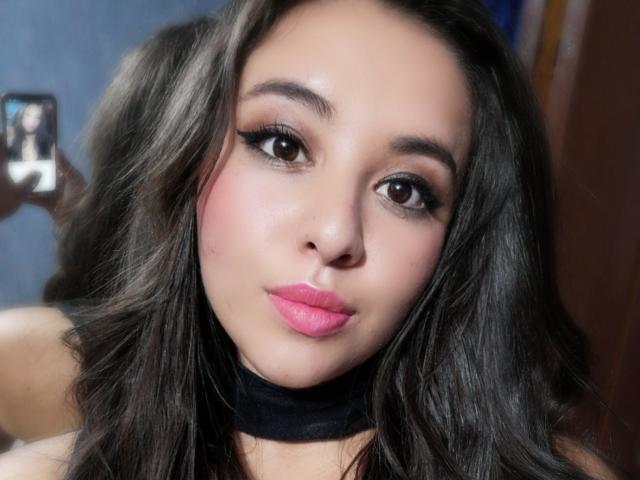 SantaRodrigues ha occhi marroni lucidi, rossetto rosa, eyeliner nero e lunghi capelli castano scuro ondulati, indossa una collana nera e ha un viso leggermente arrossato..