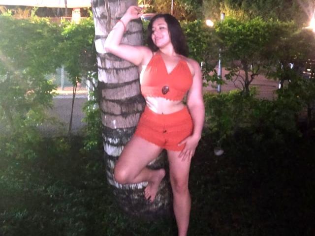 SantaRodrigues hat große Brüste, trägt ein orangefarbenes Crop-Top und Shorts, lehnt sich an einen Baum, ein Bein angehoben, lächelt, hat langes, dunkles Haar, ist barfuß und befindet sich in einem grünen Garten..