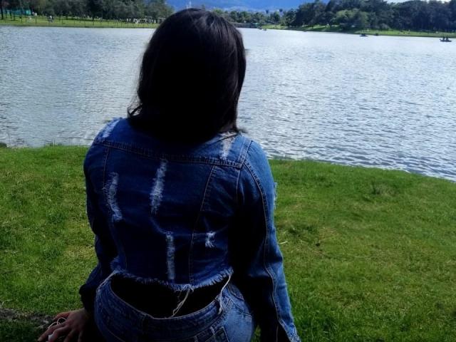 SantaRodrigues mit dunklem Haar, Jeansjacke und abgenutzten Jeans, sitzt auf dem Gras, mit Blick auf einen See und zeigt einen kleinen, schwarzen Hintern..