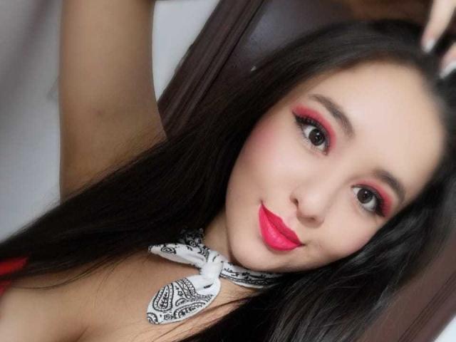 SantaRodrigues a de longs cheveux noirs, un fard à paupières rouge, un rouge à lèvres rose et une cravate blanche à motifs. Elle lève un bras avec un sourire mignon. Ses seins sont partiellement visibles, elle porte un haut rouge..