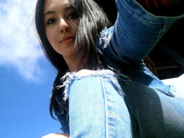 SantaRodrigues, con lunghi capelli neri, indossa jeans blu strappati, sta scattando un selfie da un'angolazione bassa, mostrando il suo viso e la parte superiore del corpo, con un cielo blu brillante sullo sfondo..