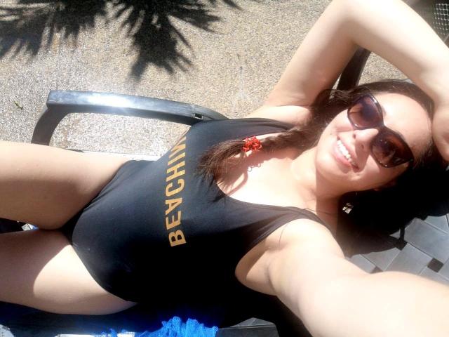 Santa Rodrigues, con i capelli castani raccolti in una treccia, indossa un costume intero nero con la scritta "BEACH" in oro, è sdraiata su una sedia, con gli occhiali da sole, sorridendo, illuminata dalla luce del sole, e ha un elastico rosso per i capelli..