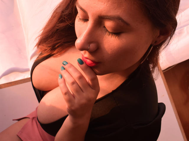 AradiaX with her eyes closed, pink lips slightly parted, touching her blue-painted nails to her chest, showing her big tits in a black top and pink skirt.