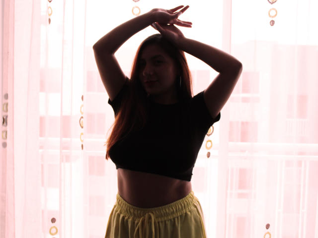 Silhouette of AradiaX with arms raised, wearing a black crop top and yellow pants, standing in front of a bright window. Her medium tits and flat stomach are visible.