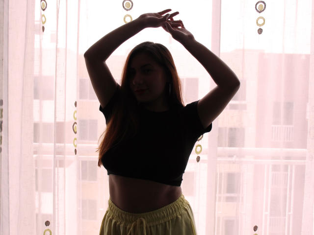 AradiaX, con la silueta delineada, con los brazos levantados, vistiendo un top corto negro y pantalones amarillos, está de pie frente a una ventana translúcida. Se pueden ver sus pechos, y tiene el pelo largo y castaño..
