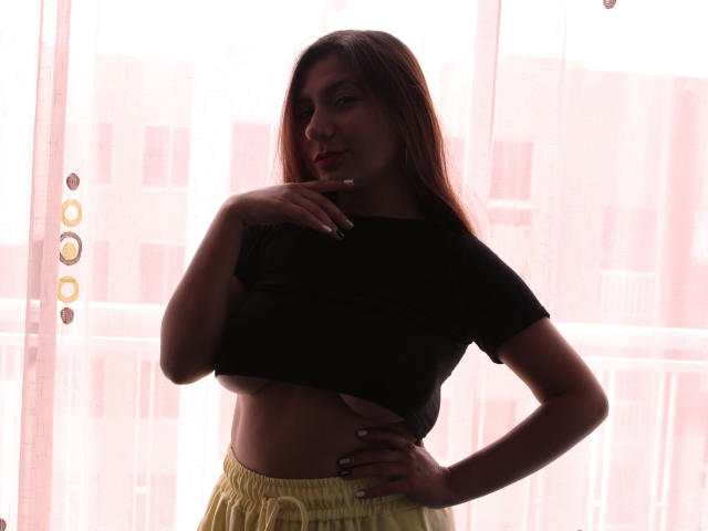 AradiaX with brown hair, black crop top, and yellow pants, holding her chin, showing her flat stomach and small tits, standing in front of a sunlit window.