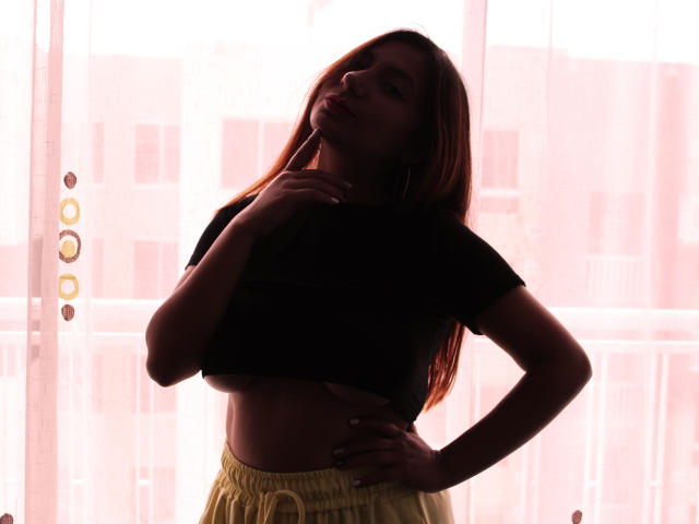 Silhouetted AradiaX with long red hair, black crop top, yellow pants, hand on hip, finger to lips, standing in front of bright window, backlit.