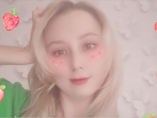 Mulher loira NaozuQAce com emojis de coração rosa nas bochechas, camisa verde, mão no cabelo, sorriso suave, lábios rosa, pele clara, fofa, ligeiramente borrada, emojis de morango nos cantos..
