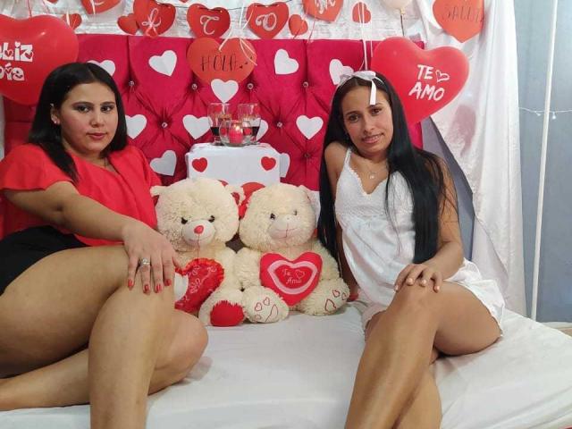 MartinaYVanessa, con cabello negro, una blusa roja y pantalones cortos negros, está sentada en la cama junto a MartinaYVanessa, que lleva un vestido blanco, y ambas muestran sus glúteos, al lado de dos ositos de peluche blancos con forma de corazón..
