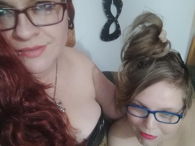 Due ragazze latine con gli occhiali, capelli rossi e biondi, e piercing al naso, che mostrano il décolleté, una con rossetto rosso e l'altra con rossetto blu. I capelli della bionda sono raccolti in uno chignon disordinato..