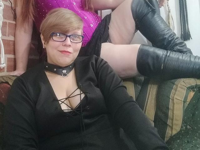 A photo of LatinoHardGirls with short blonde hair, glasses, black dress with lace-up, showing tits, and black choker, sitting next to another LatinoHardGirls in a pink sparkly top.