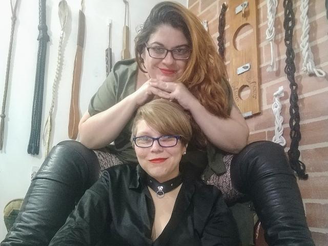 Two LatinoHardGirls in a BDSM room; one with glasses, short blonde hair, black shirt, leather boots, and collar, sitting; other with glasses, long brown hair, green shirt, holding.