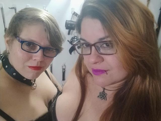 Due ragazze latine con occhiali, rossetto rosso e collari neri. Una ha i capelli castani corti, l'altra ha i capelli rossi lunghi. Entrambe hanno il seno visibile, una indossa pizzo nero..
