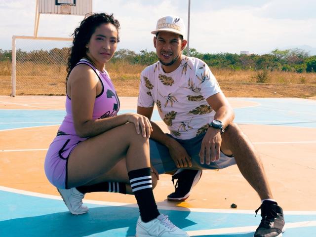 PenelopeXSmith tiene la piel bronceada, cabello negro rizado y lleva un vestido deportivo morado con calcetines negros, agachada en una cancha de baloncesto. Un hombre con un sombrero bronceado, camisa blanca y pantalones cortos está agachado a su lado..