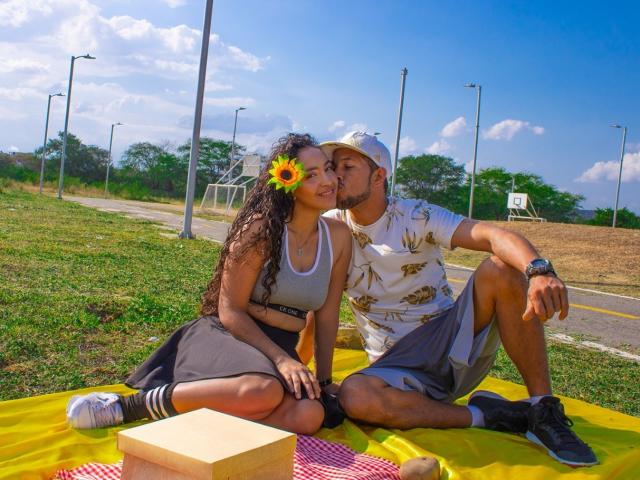 PenelopeXSmith with sunflower in hair, gray sports bra, black skirt, sitting on yellow blanket, kissing man in white shirt, gray shorts, baseball cap, yellow picnic basket, red checkered cloth.