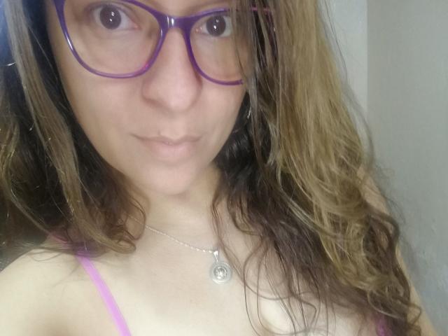 Angelonix with purple glasses, wavy brown hair, and a pink top, showing her tits and a subtle pout, wearing a silver necklace with a small circular pendant.