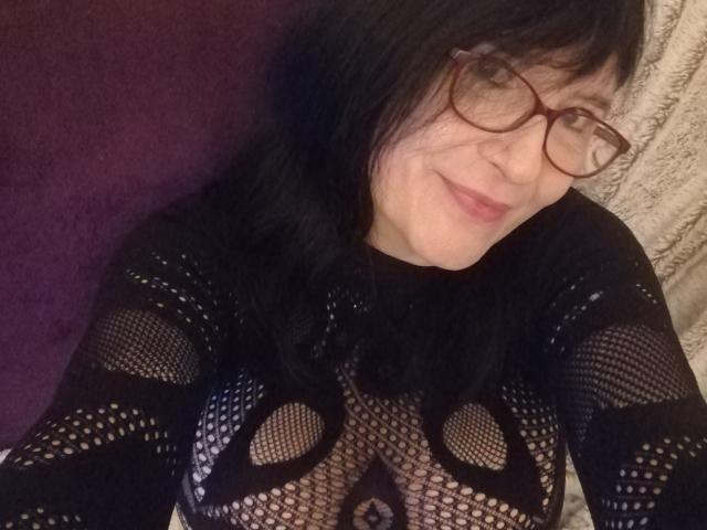 Emilianne con una blusa de malla negra que deja ver sus pechos, usando gafas, sonriendo, acostada, cabello oscuro, piel clara, un cojín morado detrás de ella y una manta beige texturizada..