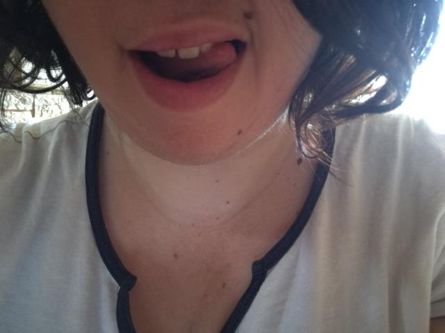 O rosto de CallieBijouFrancaise, com a boca ligeiramente aberta e a língua para fora, vestindo uma camisa branca com detalhes pretos, cabelos castanhos cacheados e uma pequena pinta na bochecha direita..