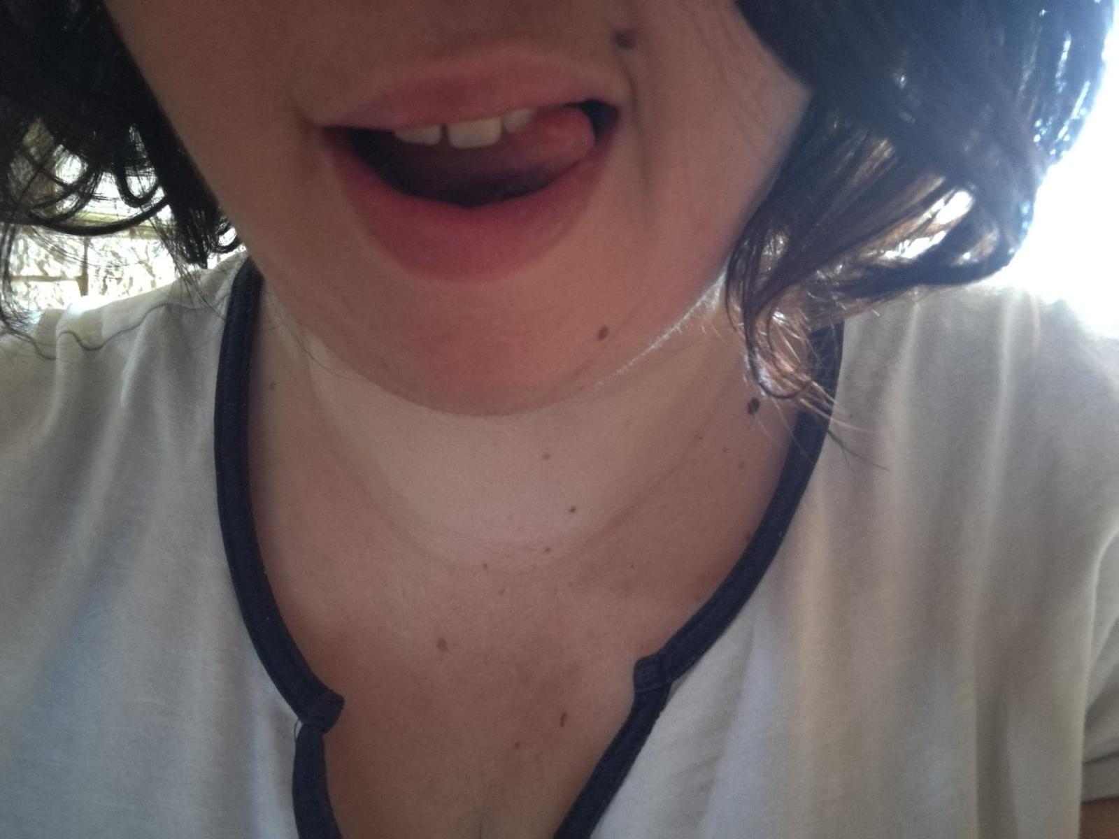 Il viso di CallieBijouFrancaise, con la bocca leggermente aperta e la lingua fuori, indossa una camicia bianca con bordi neri, ha i capelli castani ricci e una piccola lentiggine sulla guancia destra..
