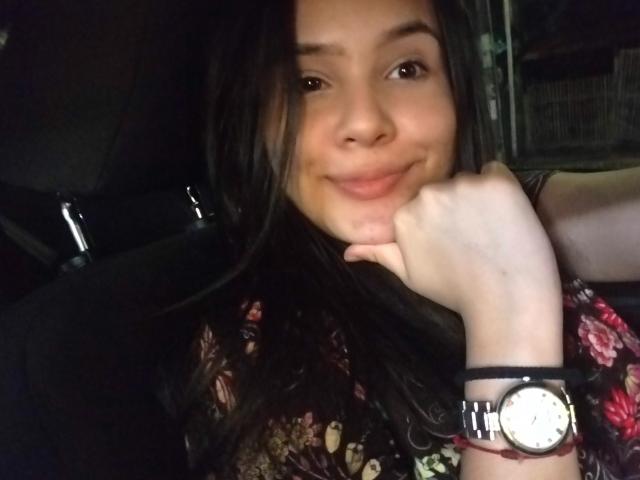 Valen, con i capelli castano scuro, sorridente, con il mento appoggiato sulla mano, indossa una maglietta a fiori e un orologio da polso, seduta in un'auto con scarsa illuminazione..