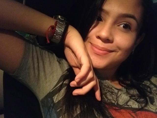 ValenDirty with dark hair, smiling, holding her long black hair with one hand, wearing a gray shirt and a silver watch with a red bracelet, in a dimly lit photo.