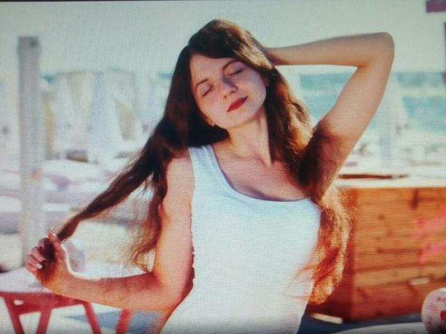 MalaniSweet, con lunghi capelli castani, occhi chiusi e un sorriso, indossa una canotta bianca, tiene i capelli sollevati con una mano, e trasmette un'aria rilassata, sensuale e sicura di sé..