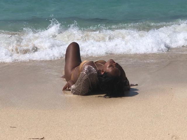 AllyCharlotee, à la peau foncée, est allongée sur une plage de sable, vêtue d'un bikini marron, les jambes pliées, la tête reposant sur le sable, de l'eau éclabousse autour d'elle, ses cheveux sont partiellement mouillés..