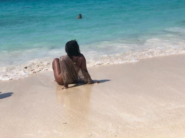 AllyCharlotee, sitting on a sandy beach with her ass down, wearing a netted top, looking at the ocean, with a person's head in the water in the background.