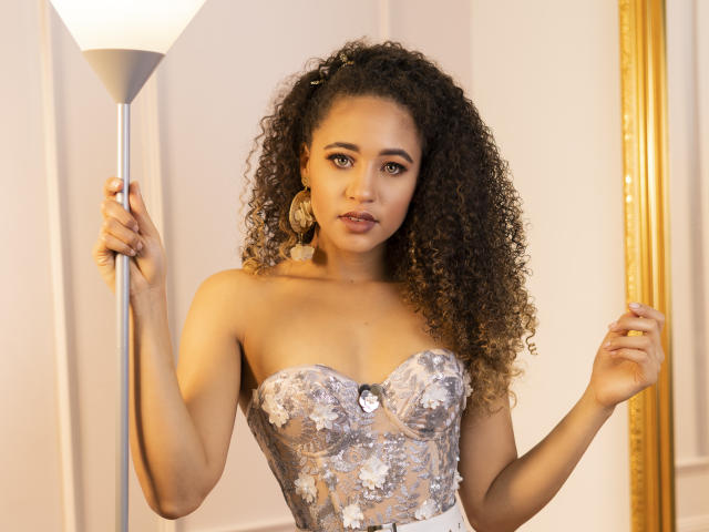 AlyssaBelle with curly brown hair, wearing a strapless silver dress with floral lace, holding a lamp, looking at the camera with a slight smile, showing her tits.