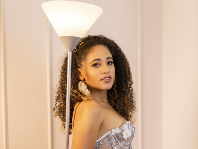 AlyssaBelle mit lockigem, braunem Haar, heller Haut und kleinen Brüsten, trägt ein trägerloses, gemustertes Oberteil und steht neben einer weißen Stehlampe, während sie in die Kamera schaut..