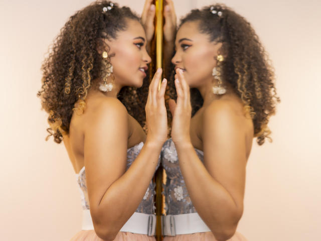 AlyssaBelle, avec ses cheveux bouclés, portant une robe fleurie sans bretelles, se tient devant un miroir, les mains jointes, reflétant son profil et son expression coquette..