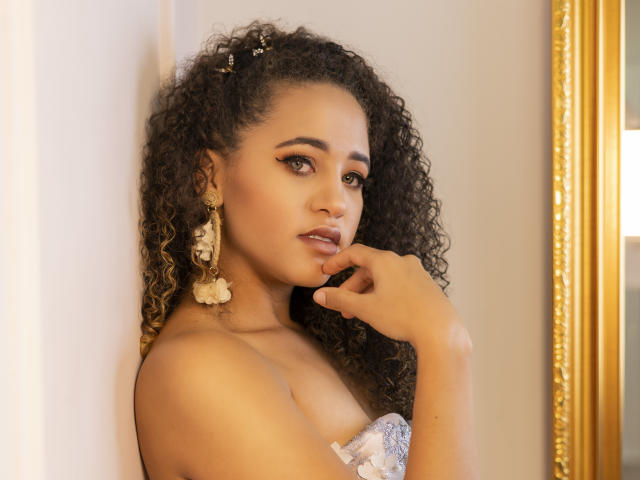 AlyssaBelle, met krullend bruin haar, gouden oorbellen en een top zonder bandjes, die haar lippen aanraakt met een verleidelijke blik, waarbij haar gladde, bruine huid en subtiele make-up te zien zijn..