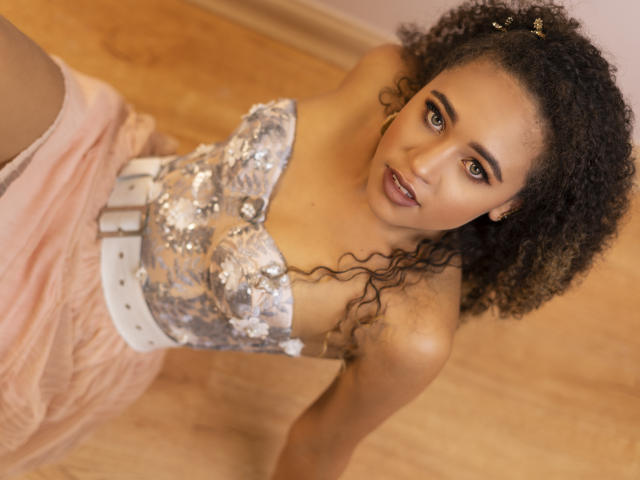 AlyssaBelle, avec des cheveux bouclés, portant un haut argenté pailleté et une jupe rose, regardant vers le haut avec une expression séduisante, ceinture blanche, sol en bois, éclairage chaleureux et maquillage léger..