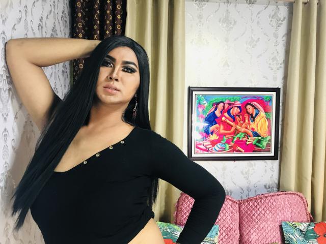 Née avec de longs cheveux noirs, portant un haut noir à une épaule, une main dans les cheveux, regardant la caméra, exhibant sa poitrine et affichant une expression confiante et sexy..