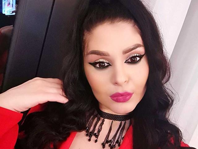 LexyRene heeft donker, golvend haar, opvallende zwarte eyeliner, roze lippenstift, een zwarte choker met kralen en een rode top. Haar hand is in de buurt van haar hoofd..