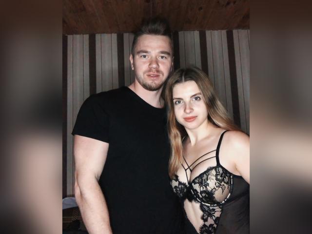 HotLoversVida mit mittelgroßer Brust, in schwarzer Spitzenunterwäsche, steht neben HotLoversVida mit kurzen Haaren, schwarzem T-Shirt und leichten Bartstoppeln. Beide haben helle Haut und lächeln..