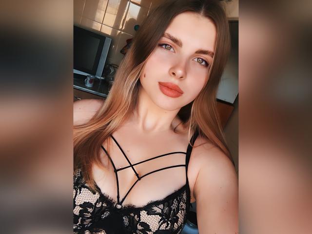 HotLoversVida with light brown hair, blue eyes, and orange lipstick, wearing a black lace bra with strappy details, showing her tits, in a sunlit room.