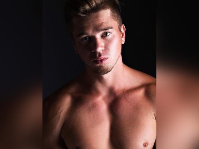 HotLoversVida, senza camicia, con un petto tonico e muscoloso, capelli castani corti e una leggera barba, ha un'espressione seria ed è parzialmente illuminato in un ambiente buio..