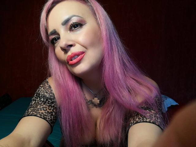 HotAdelayde69, avec ses cheveux roses, son haut à motifs léopard, ses lèvres rouges et son collier avec une croix, sourit à la caméra et montre ses seins. Elle a un maquillage des yeux foncé et un tatouage mignon..