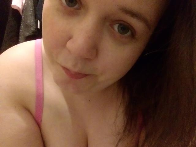 LoveClo heeft bleke huid, bruine ogen en roze beugels van haar bh, die haar borsten laten zien in een selfie met een lichte, onschuldige glimlach, en bruin haar dat op haar rechter schouder valt..