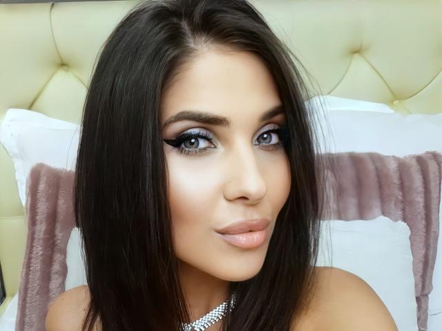 LovelyKinsley has dark, straight hair, blue eyes with heavy eyeliner, and a slight smile. She's wearing a silver necklace and has pink, glossy lips.