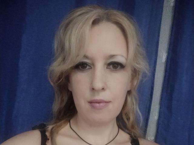 SoftGella tiene el cabello rubio y ondulado, delineador de ojos negro y lápiz labial rosa. Lleva una blusa negra de encaje con un collar negro. Sus glúteos no son visibles en esta imagen..