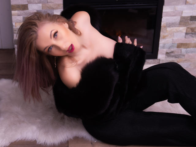 LadyLea with pink lips, blue eyes, and brown hair, sitting on a white rug, wearing a black fur off-shoulder coat and black pants, exposing her tits.
