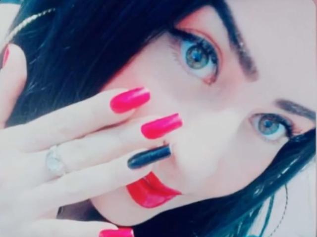 LindaSmith's face with blue eyes, black eyebrows, red lipstick, and long black nails, featuring one black nail and a silver ring on her left hand.